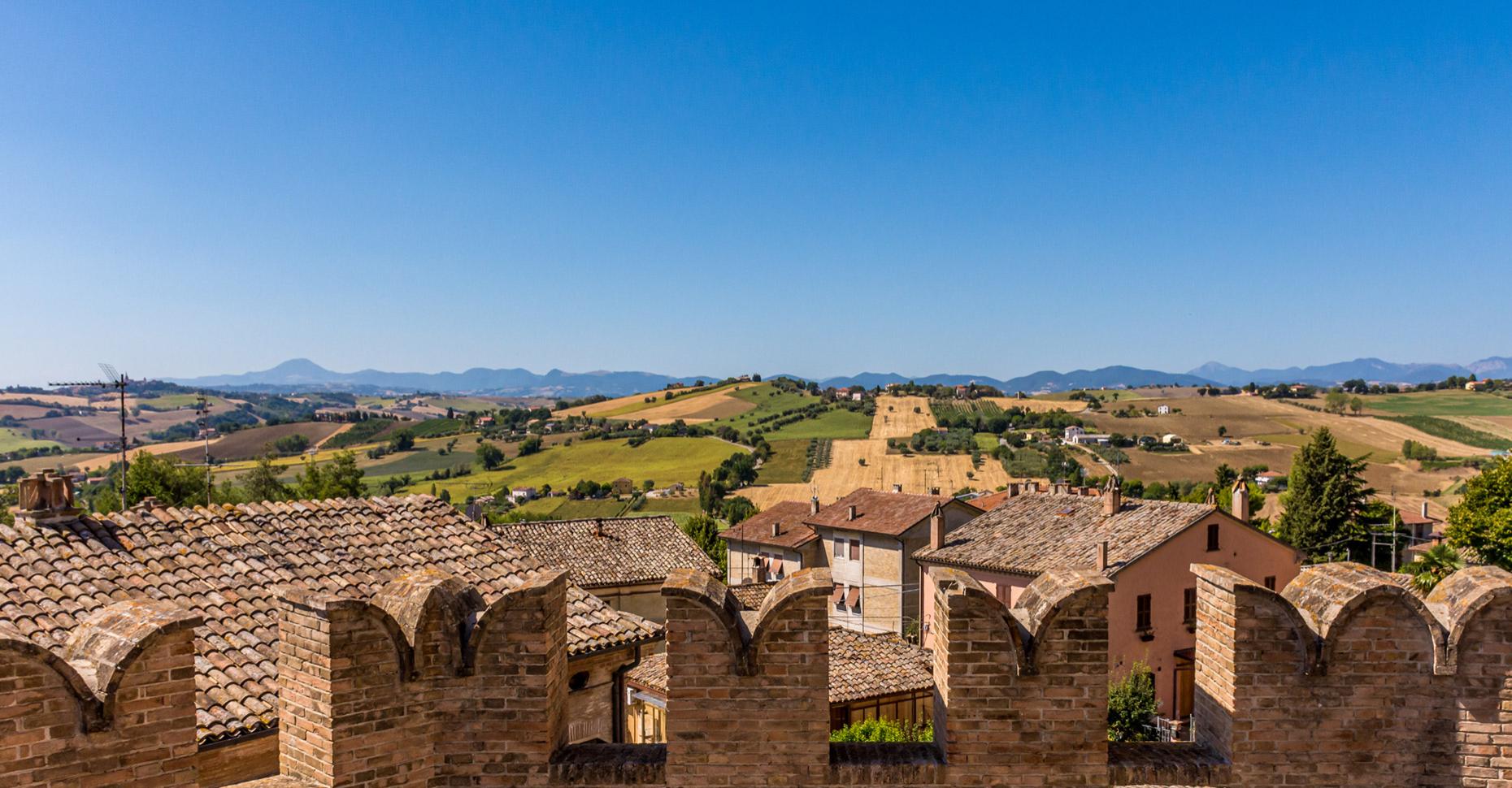 Paesaggio collinare con campi coltivati e tetti di case sotto un cielo azzurro.