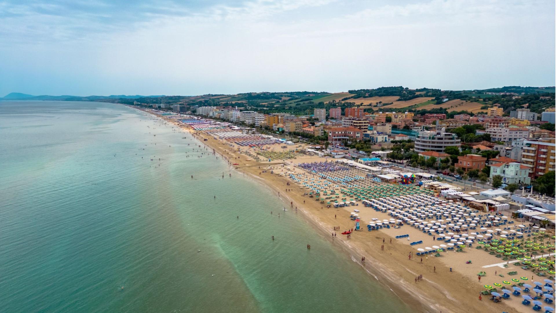 Spiaggia affollata con ombrelloni colorati e mare calmo, città costiera sullo sfondo.