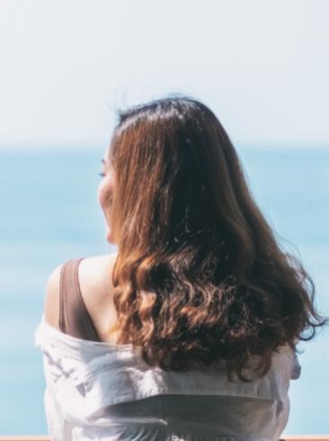 Donna di spalle, capelli mossi, guarda il mare in una giornata soleggiata.