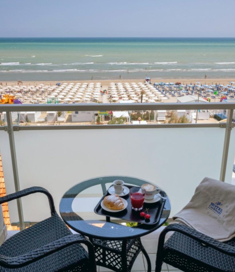 Colazione sul balcone con vista mare, spiaggia e ombrelloni, atmosfera rilassante.