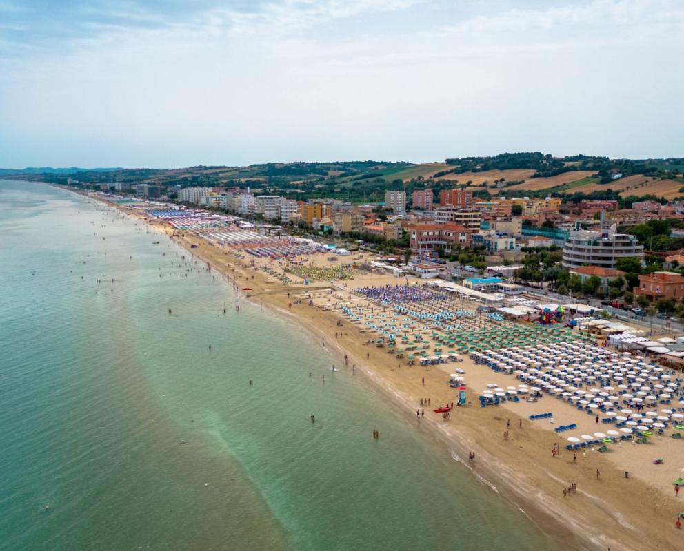 Spiaggia affollata con ombrelloni colorati e mare calmo, città costiera sullo sfondo.